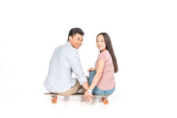 cheerful asian couple sitting on longboard and smiling at camera on white background - Foto, afbeelding