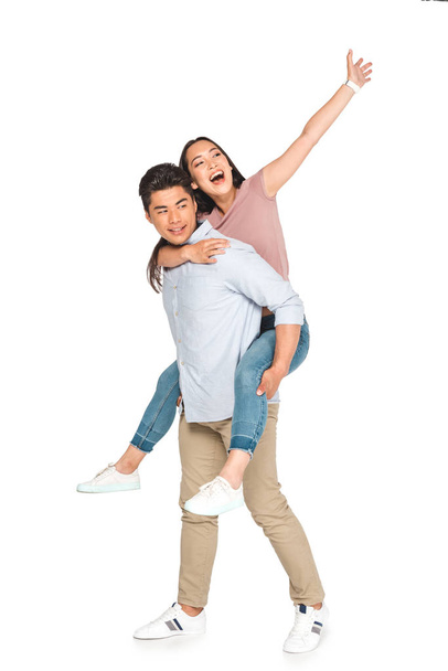 cheerful asian woman waving hand while piggybacking on happy boyfriend on white background - 写真・画像
