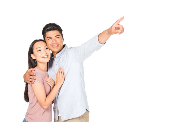 happy asian man hugging smiling girlfriend while pointing with finger isolated on white - Fotó, kép