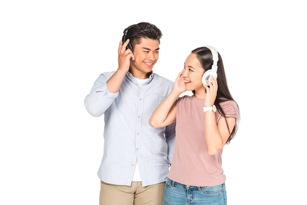cheerful asian couple looking at each other while listening music in headphones isolated on white - Foto, imagen