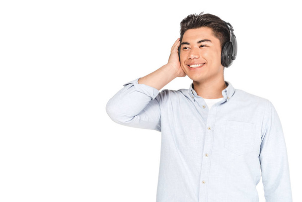 Wesoła Asian Man słuchania muzyki w słuchawkach, uśmiechając się i patrząc z dala na białym tle - Zdjęcie, obraz
