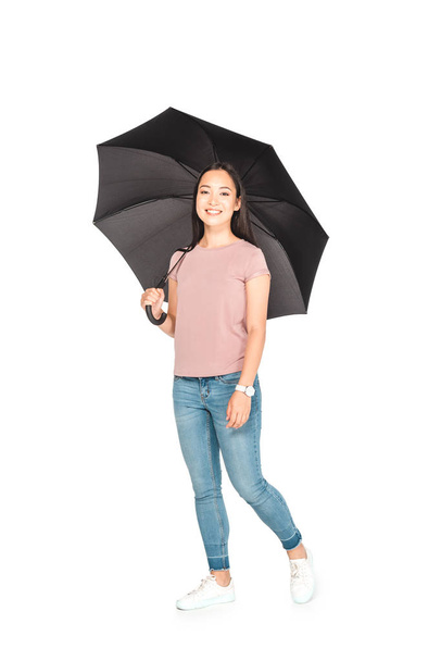 cheerful asian girl holding black umbrella and smiling at camera on white background - Photo, Image