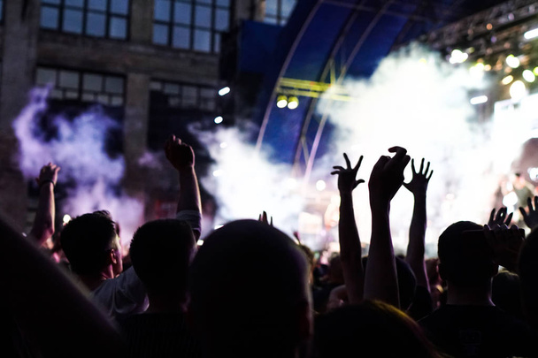 Концертная толпа, посещающая концерт, видны силуэты людей, подсвеченные огнями сцены. Поднятые руки и смартфоны видны здесь и там
. - Фото, изображение
