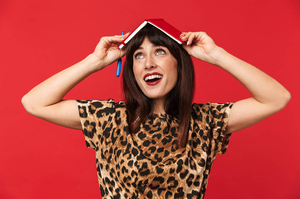 Woman dressed in animal printed shirt posing isolated over red background with notebook on head. - Foto, imagen
