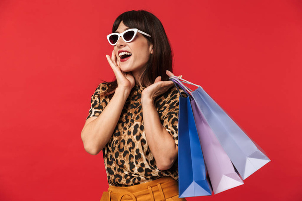 Beautiful excited young woman dressed in animal printed shirt posing isolated over red background wearing sunglasses holding shopping bags. - Фото, изображение