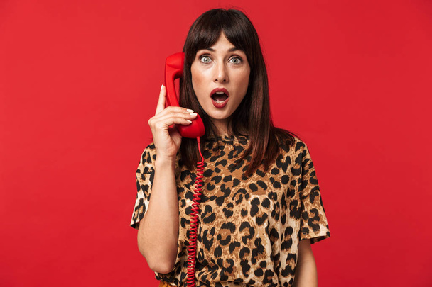Shocked young woman dressed in animal printed shirt posing isolated over red background talking by telephone. - Fotografie, Obrázek