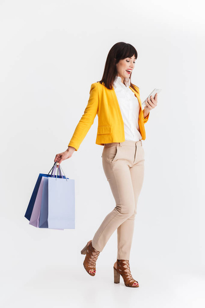 Beautiful young business woman posing isolated over white background using mobile phone holding shopping bags. - Foto, imagen