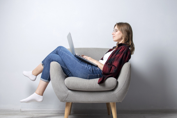 Młoda dziewczyna z laptopem siedzi na miękkim wygodnym fotelu i sfrezowanie, Kobieta za pomocą komputera przed białą pustą ścianę, ona Freelancing i drukowanie tekstu, Kopiuj przestrzeń - Zdjęcie, obraz