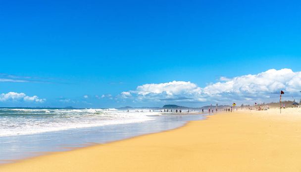 wunderschöner weiträumiger Panoramablick auf die Wellen des Ozeans, die in Richtung tropischer Sandstrand rollen und Silhouetten von Surfern und Strandbesuchern, die sich in der Ferne entspannen. - Foto, Bild