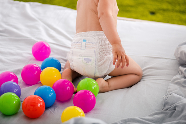 Cropped view of cute baby in diaper sitting on bed among colored balls - Foto, imagen