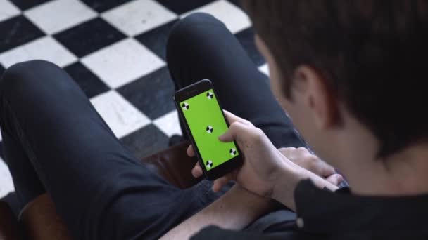 Pohled z ramene muže v černé košili a džínách, které drží smartphone v ruce a poklepávat na obrazovku prstem v režimu na výšku se zelenou obrazovkou. Skladní záběry. Zelená obrazovka klíče Chroma - Záběry, video
