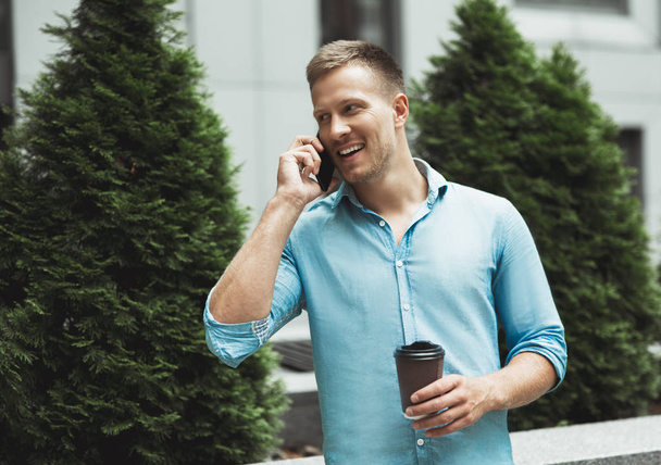 young handsome smiling man drinks coffee on the go standing near office business center speaking on his smartphone - 写真・画像