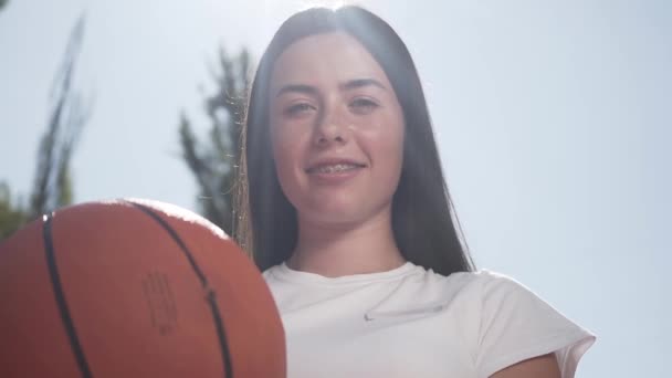 Portrait of attractive brunette woman with a braces holding basketball ball looking at the camera smiling. Concept of sport, power, competition, active lifestyle. Girl playing basketball on the court - Záběry, video