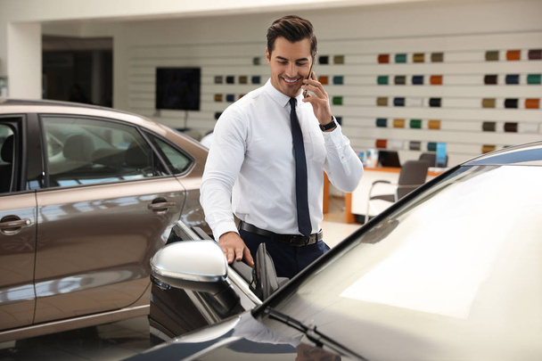 Young man talking on phone near car in modern dealership - Zdjęcie, obraz