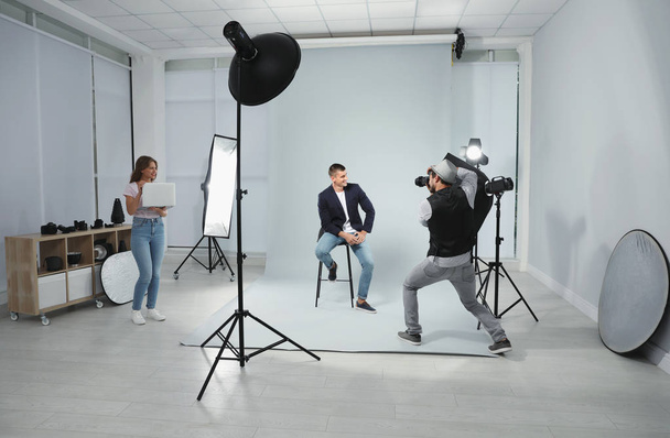 Professional photographer with assistant taking picture of young man in modern studio - Фото, зображення