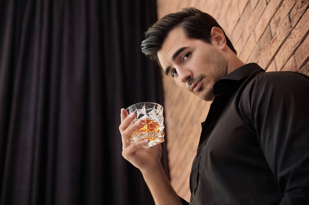 Young man with glass of whiskey near brick wall indoors. Space for text - Fotoğraf, Görsel