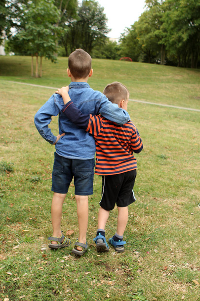boys, preschooler, primary school student are standing with their backs together, hugging in the park - Valokuva, kuva