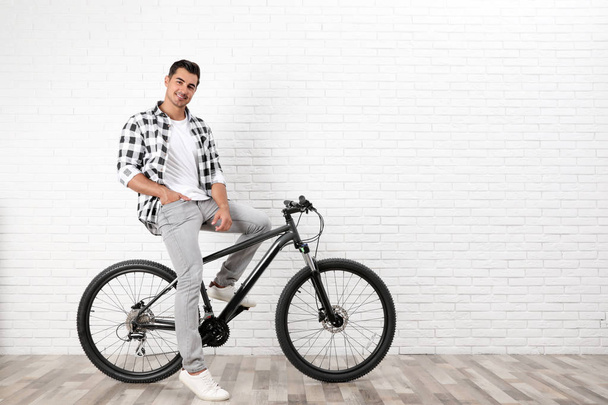 Handsome young man with modern bicycle near white brick wall indoors - Foto, imagen