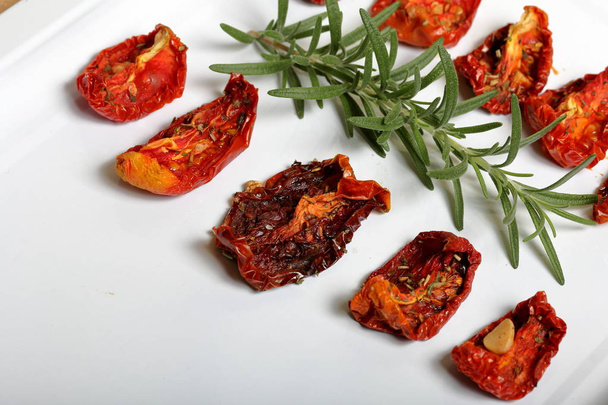 Sun-dried tomatoes with spices and garlic on a light tray. Nearby lies fresh rosemary. - Foto, Imagen