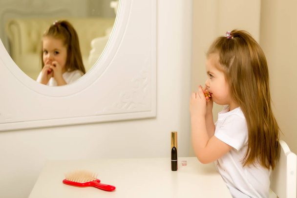 A little girl near the mirror paints lipstick lips - Φωτογραφία, εικόνα