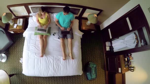 ARRIBA: Joven pareja caucásica de vacaciones en Vietnam trabajando en sus computadoras portátiles mientras están sentados en la cama king size. Turistas femeninos y masculinos navegando por Internet desde su cómoda habitación de hotel
. - Imágenes, Vídeo