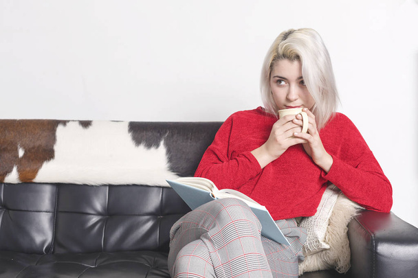 Caucasian blonde woman sitting on sofa drinking coffee and reading book - Foto, immagini