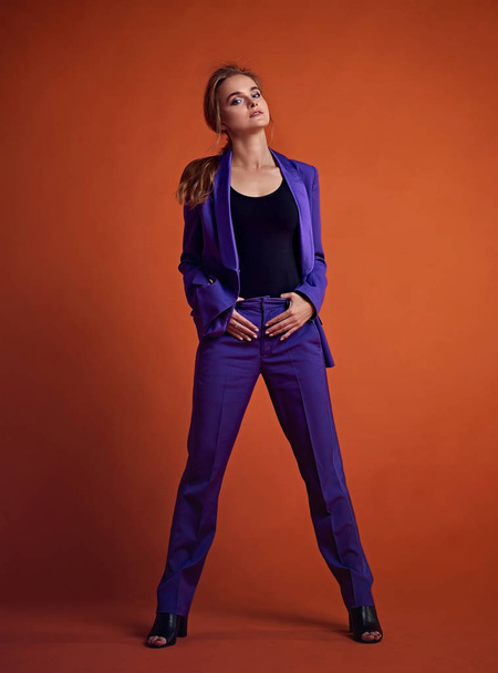 若い魅力的な女性モデルは、紫色のスーツを着て、スタジオでポーズ。オレンジ色の背景. - 写真・画像
