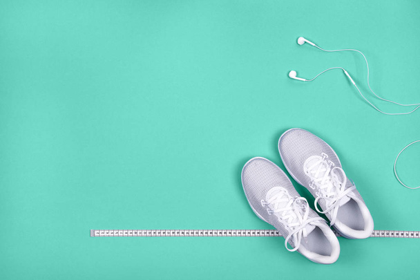Top view of white sport shoes (sneakers) with measuring tape and white earphones (headphones) on blue-green background. - Photo, Image
