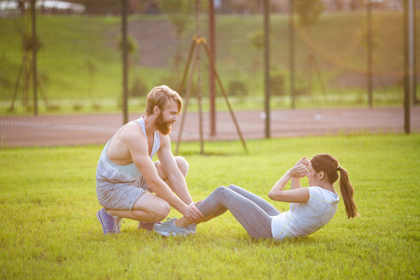 Siedzieć UPS fitness para ćwiczenia siedzieć na zewnątrz w trawie. Dopasowanie osób pracujących na trening przekrojowy. Kobieta robi brzuch brzuszków prasy ćwiczenia z trenerem. Para robi Sit-UPS brzucha Crunch - Zdjęcie, obraz