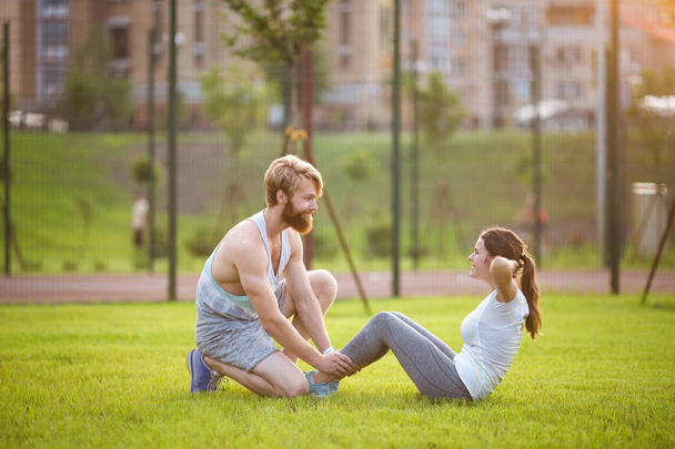 Sit ups fitness couple exercising sit up outside in grass. Fit people working out cross training. Woman doing abdominal crunches press exercise with trainer. Couple Doing Sit-Ups Abdominal Crunch - Photo, Image