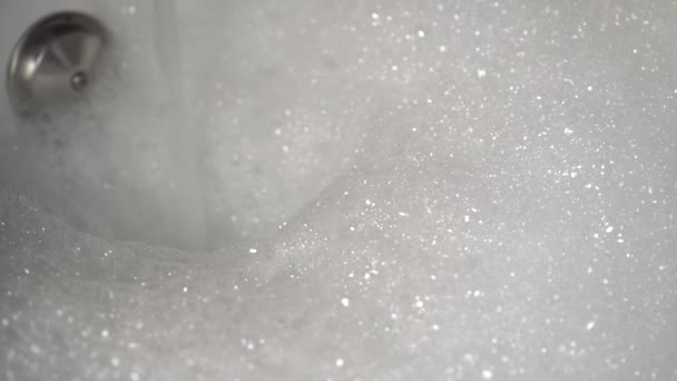 Una corriente de agua vierte en el baño y hace espuma del producto de higiene. Una pila de espuma está creciendo. Preparación para lavado corporal
 - Imágenes, Vídeo
