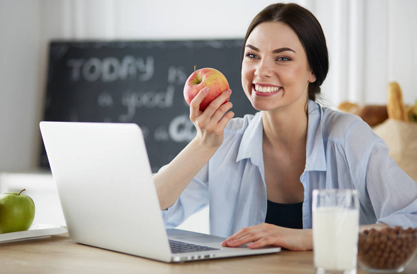 Nainen omena ja kannettava tietokone istuu keittiössä
 - Valokuva, kuva