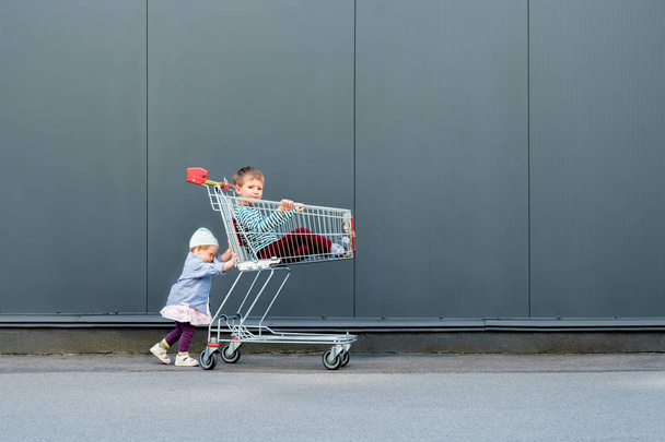 Two trendy hipsters with shopping cart near supermarket, shopping mall. Two cute children waiting for parents outdoor. Happy childhood concept background. Little girl playing with her brother - Photo, Image