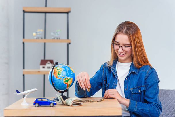 Beautiful caucasian girl counts her savings to foreign vacation. Young woman sits at table with glass jar, money, globe, model of air plane and car. - 写真・画像