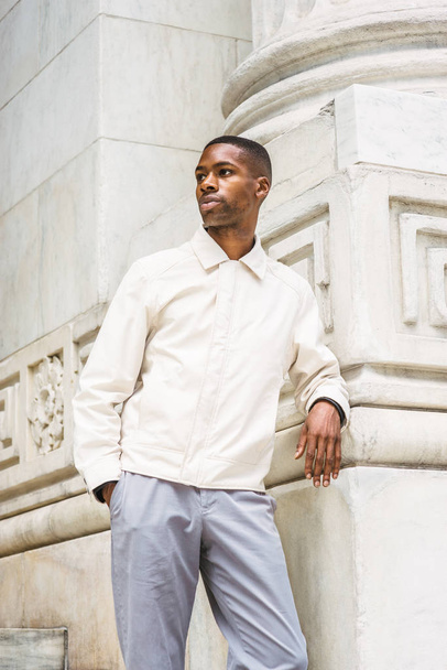Portrait of Young Handsome African American Man in New York. Young black man wearing light color jacket, standing by vintage marble wall, looking around, relaxing,  thinking - Photo, Image