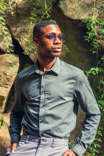 Fiatal jóképű African American Man viselése szürke inget, napszemüveget, állva sziklák fal alatt a nap, körülnézett, pihentető a Central Park, New York - Fotó, kép