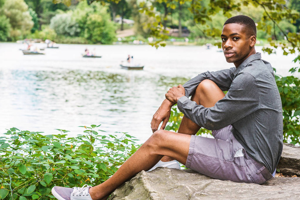 Young African American Man relaxing at Central Park in New York. Young black man wearing long sleeve gray shirt, shorts, sitting on rocks by lake, looking around. People rowing boats on background - Foto, afbeelding