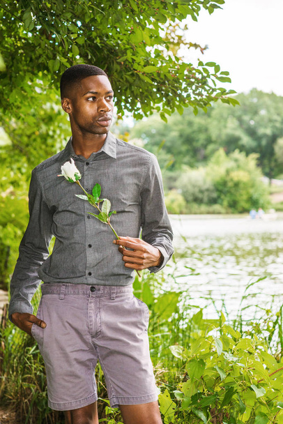 Młody African American Man brakuje Ci i czeka na Ciebie w Central Park w Nowym Jorku. Młody czarny człowiek stoi nad jeziorem, trzymając białą różę, czekając, myśląc, stracił w myśleniu. - Zdjęcie, obraz