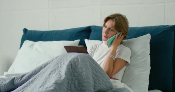 Young woman lying in bed, talking on her smartphone, using gadgets - Materiał filmowy, wideo
