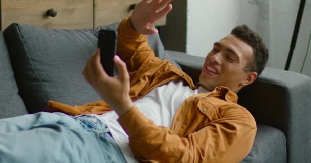 Hispanic man is lying on sofa, having a video chat via smartphone - Video
