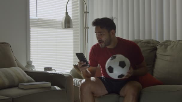 Happy man at home watching soccer game on cellphone while holding soccer ball  - Filmagem, Vídeo
