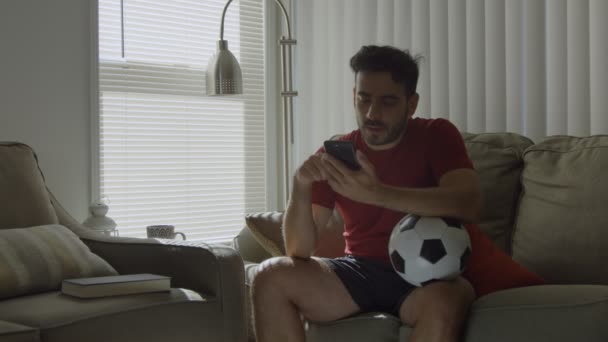 Soccer fan checking social media while holding soccer ball at home - Záběry, video