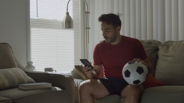 Man watching soccer game on cellphone while holding soccer ball at home - Filmagem, Vídeo