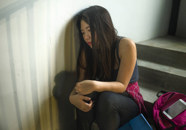 Dramatic portrait of Asian female college student bullied. Young depressed and sad Chinese girl sitting lonely on campus staircase suffering bullying and harassment - Foto, Bild