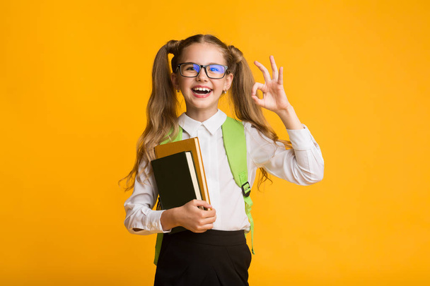 Adorable First-Grade Schoolgirl Gesturing OK At Camera On Yellow Background - Φωτογραφία, εικόνα