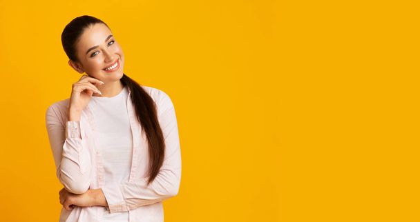 黄色のスタジオの背景の上にあごに触れるカメラで微笑む女性 - 写真・画像