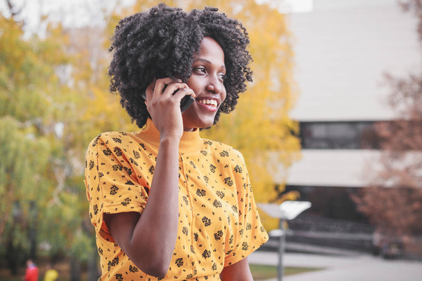 junge lächelnde dunkelhäutige Frau in lässiger Kleidung im Gespräch mit ihrem Handy - Foto, Bild