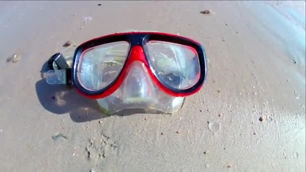 Egy Búvármaszk fekszik a homokos parton, a tenger hullámai szörföznek rajta egy napsütéses napon. Lassú mozgás standard mozgás. - Felvétel, videó