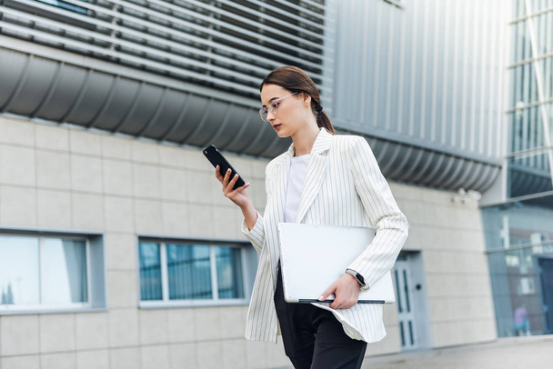 Beautiful young businesswoman wearing white shirt and using modern smart phone while walking at break in the city, professional female employer typing text message on cellphone outside - Φωτογραφία, εικόνα