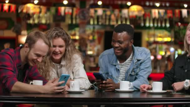 Cuatro amigos sentados en un café ríen y sonríen mientras miran las pantallas de los teléfonos e intercambian fotografías de viajes. Afro-americana novia charlando y bebiendo café
. - Metraje, vídeo